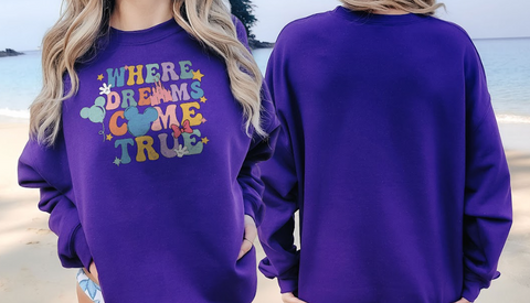 A woman wearing a purple sweatshirt with the text "WHERE DREAMS COME TRUE" printed on it, standing on a beach.