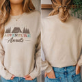 A woman wearing a beige sweatshirt with the text "Adventure Awaits" printed on it, standing in front of a plant.