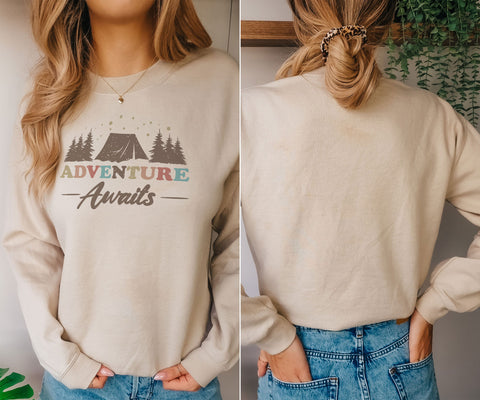 A woman wearing a beige sweatshirt with the text "Adventure Awaits" printed on it, standing in front of a plant.