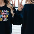 A woman wearing a black sweatshirt with a colorful graphic design and the text "WHERE DREAMS COME TRUE" is standing on a beach.
