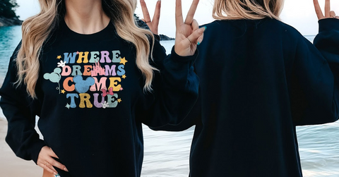 A woman wearing a black sweatshirt with a colorful graphic design and the text "WHERE DREAMS COME TRUE" is standing on a beach.