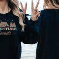 A woman with long blonde hair is standing on a beach, wearing a black sweatshirt with the text "Adventure Awaits" printed on it.