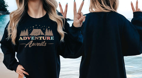 A woman with long blonde hair is standing on a beach, wearing a black sweatshirt with the text "Adventure Awaits" printed on it.