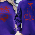 A person wearing a purple sweatshirt with a red graphic design on the front and back, standing on a beach.