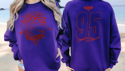 A person wearing a purple sweatshirt with a red graphic design on the front and back, standing on a beach.