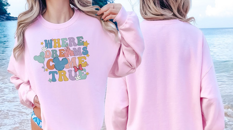 A woman wearing a pink sweatshirt with the text "WHERE DREAMS COME TRUE" printed on it, standing near the ocean.