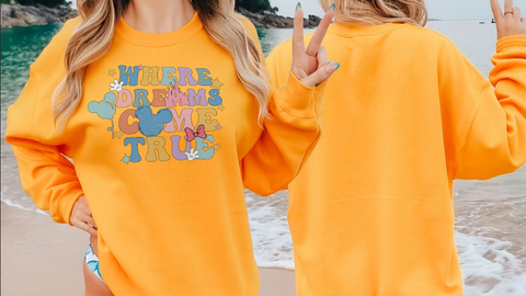 A woman wearing a bright yellow sweatshirt with the text "WHERE DREAMS COME TRUE" printed on it, standing on a beach with the ocean in the background.